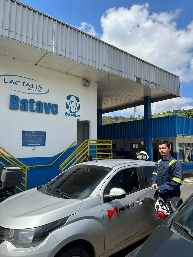 Profissional da Máximo Engenharia uniformizado e com capacete, ao lado do veículo da empresa na entrada de uma indústria.